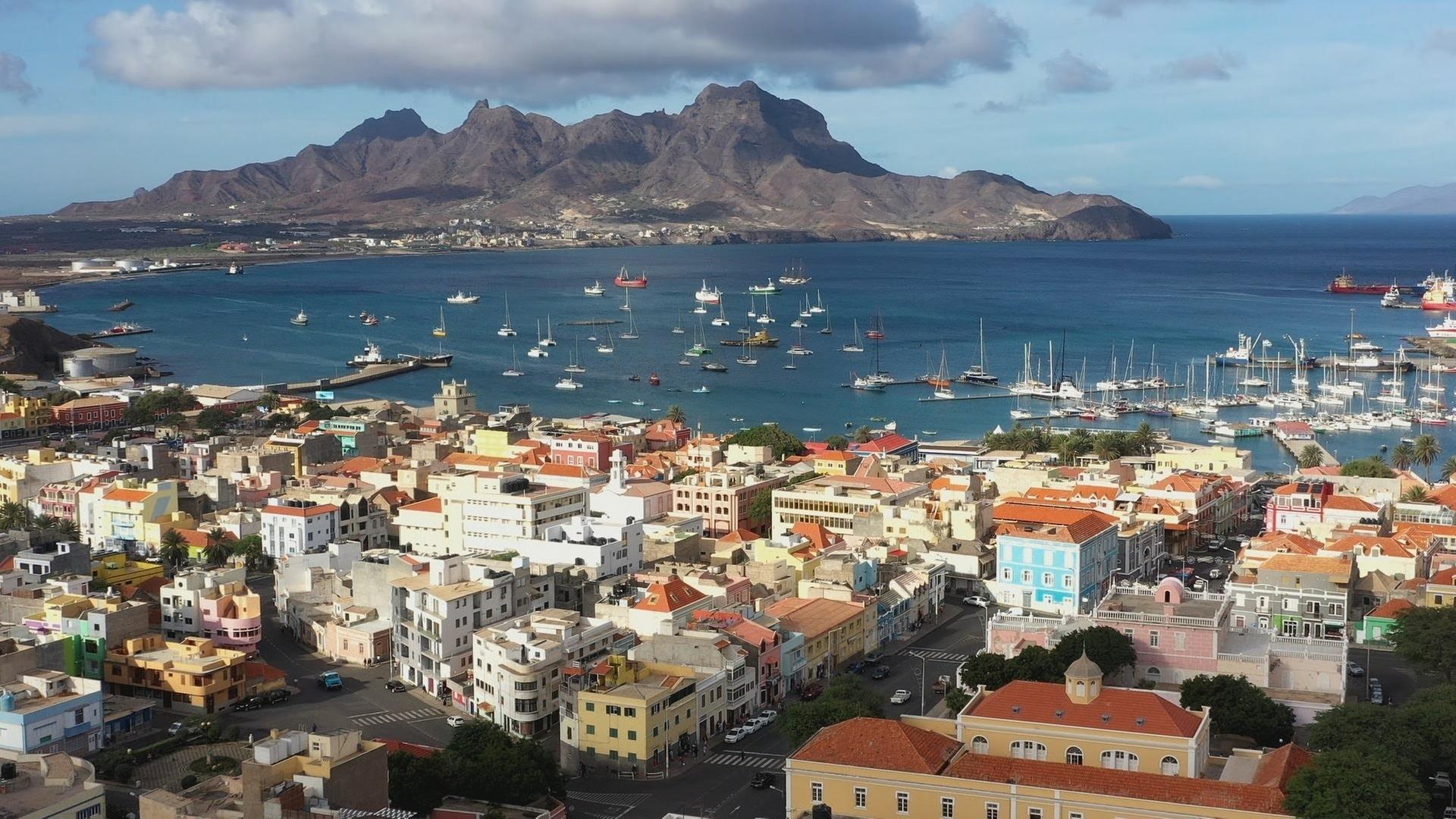 Aufnahme der Hafenstadt Mindelo mit bunten Häusern, in der Bucht und im Hafen davor liegen zahlreiche Boote. Im Hintergrund ragt ein Berg auf.