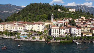 Blick vom See aus auf eine mittelalterliche Stadt mit weißen und gelben Häusern und einem Kirchturm. Dahinter erhebt sich ein dicht bewachsener Hügel.