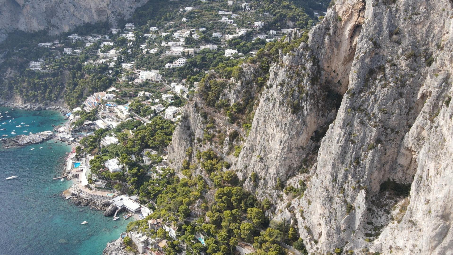ine zum Teil bewachsene Steilküste, im Hintergrund liegt eine kleine Bucht mit einer Ortschaft.