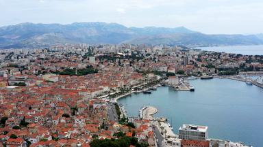 Luftaufnahme der Hafenstadt Split. Auf einer Seite stehen dicht gedrängt Häuser, auf der anderen Seite erstreckt sich das Meer. In der Mitte liegen die Hafenanlagen und die Promenade.