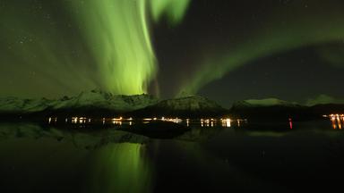 Nordlichter über schneebedeckten Bergen. Im Vordergrund beleuchtete Häuser am Meer.