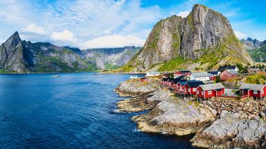 Blick auf das Küstendorf Hamnøy auf den Lofoten mit roten Fischerhäusern auf felsigem Ufer, blauem Meer und steil aufragenden Bergen im Hintergrund.