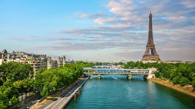 Blick über die Seine in Paris mit einer Brücke und grünen Uferpromenaden. Im Hintergrund ragt der Eiffelturm vor blauem, leicht bewölktem Himmel auf.