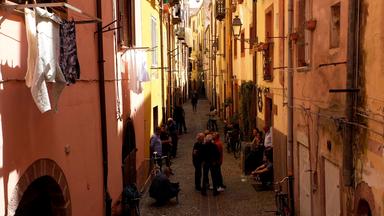 Enge Altstadtgasse im Dorf Bosa mit ockergelben Häuserwänden und einigen Menschen.