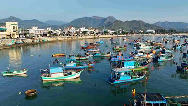 Bunte, meist blaue Boote liegen an der Küste vor Anker. Im Hintergrund Berge
