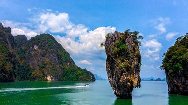 Steile, dicht bewachsene Kalksteinfelsen ragen aus dem grün-türkisen Wasser vor der James-Bond-Insel in Thailand. Ein Boot fährt zwischen den Felsen hindurch.