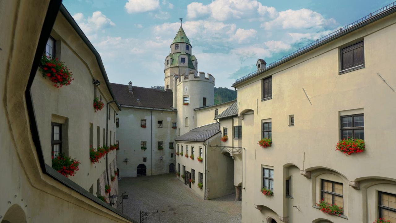 Eine Burganlage in einem Innenhof mit Blumen geschmückten Fenstern, im Hintergrund ein Turm mit grüner Kuppel.
