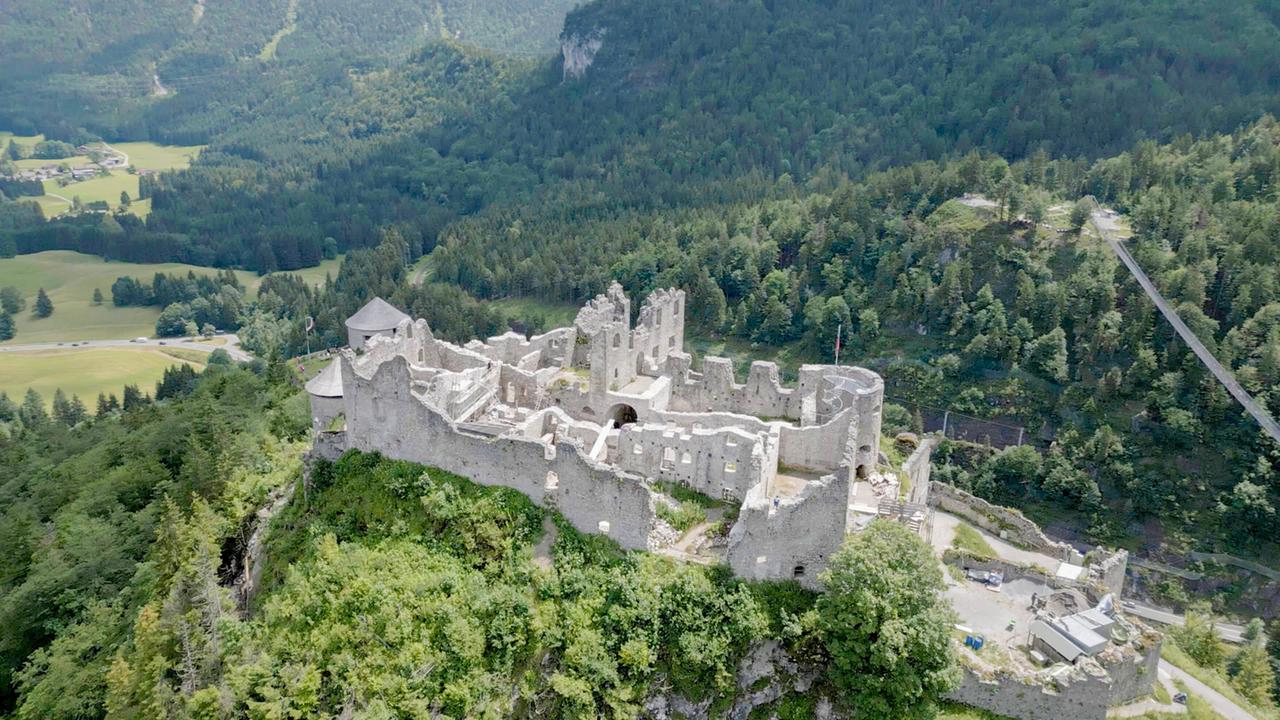 Eine Burgruine auf einem bewaldeten Hügel, umgeben von grünen Bäumen und mit Blick auf eine umliegende Landschaft.