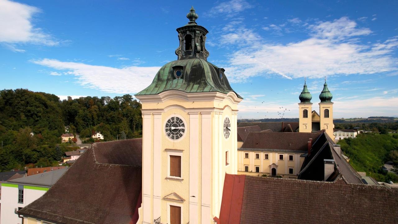 Das Bild zeigt den Kirchturm eines Klosters, wahrscheinlich das Stift Lambach in Österreich. Der Turm hat eine markante, grün patinierte Kupferkuppel und zwei kleinere Türme mit glockenförmigen Dächern, die ebenfalls grün sind. Auf der Vorderseite des Hauptturms befindet sich eine große Uhr mit drei Ziffernblättern, jeweils mit schwarzen Ziffern auf weißem Grund. Der Hintergrund zeigt einen klaren blauen Himmel mit wenigen Wolken. Rund um das Kloster ist eine grüne, bewaldete Landschaft zu sehen. Im Vordergrund sind Dächer von angrenzenden Gebäuden zu erkennen, deren Materialien und Farben variieren. Die Atmosphäre wirkt ruhig und historisch.