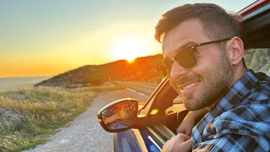 Ein junger Mann streckt den Kopf aus dem Fahrerfenster eines Wagens auf einer kleinen Straße.