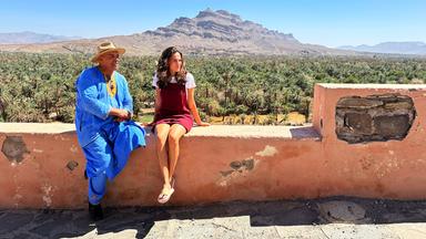 Eine junge Frau und ein Mann sitzen auf einer Mauer über einer Oase.