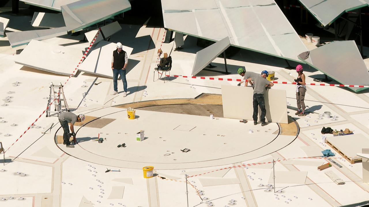 Das Bild zeigt eine Szene vom Aufbau eines Bühnenbildes für die Bregenzer Festspiele. Auf einer großen, hellen Fläche sind mehrere Personen mit Helmen und Arbeitskleidung zu sehen, die an verschiedenen Aufgaben arbeiten.   In der Mitte des Bildes ist ein kreisförmiges Element, das teilweise aus Holz besteht. Rund um dieses Element befinden sich zahlreiche kleine, runde Objekte, die wahrscheinlich zur Befestigung dienen. Ein Arbeiter beugt sich über das kreisförmige Element, während ein anderer im Hintergrund an einem großen, vertikalen Stück arbeitet.  Im Hintergrund sind größere, schräg angeordnete Teile zu sehen, die an das Bühnenbild angehängt werden. Rote und weiße Absperrbänder sind an verschiedenen Stellen angebracht, um den Arbeitsbereich abzugrenzen. Der Himmel scheint klar und sonnig zu sein, was auf eine angenehme Wetterlage hinweist.   Das Bild vermittelt den Eindruck von geschäftiger Aktivität und Vorfreude auf die bevorstehende Aufführung.