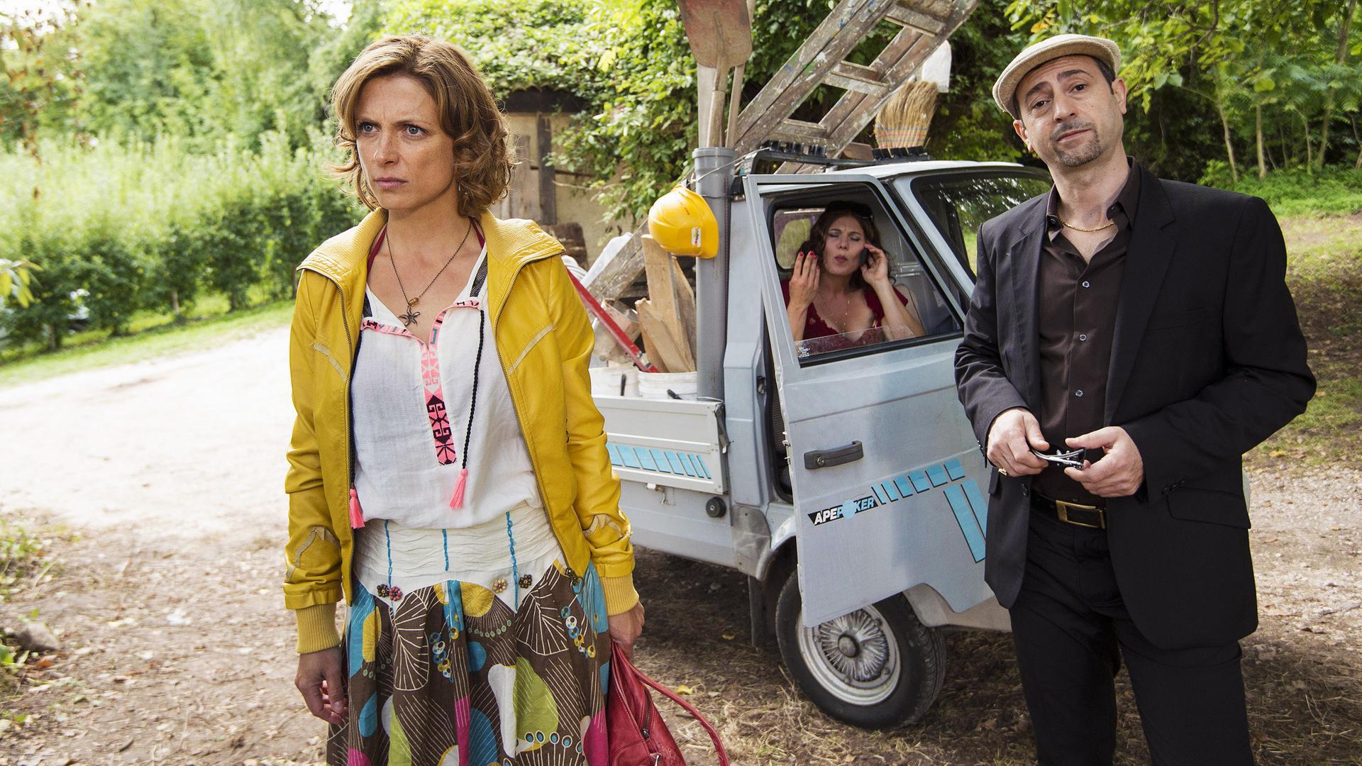 Eva (Julia Brendler, vorne) blickt genervt. Neben ihr steht der italienische Handwerker Giovanni im Anzug. Im Hintergrund sitzt seine Verlobte Isabella (Yasmina Djaballah) im Auto.