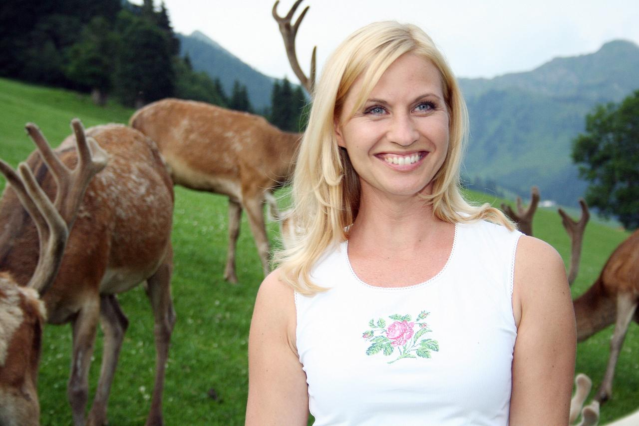 Das Bild zeigt eine blonde Frau mit langen Haaren, die in einem weißen Top mit floralem Muster am Ausschnitt gekleidet ist. Sie lächelt freundlich in die Kamera. Im Hintergrund sind mehrere Rehe zu sehen, die auf einer grünen Wiese grasen. Die Wiese wird von Bergen umgeben, die teilweise bewaldet sind. Der Himmel ist bewölkt, was darauf hinweist, dass es ein bewölkter Tag ist. Das Gesamtbild vermittelt eine harmonische, naturnahe Atmosphäre.