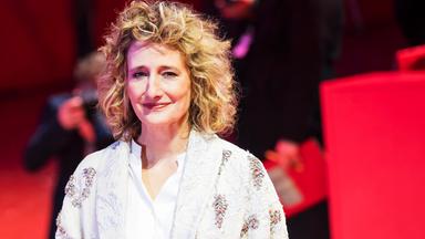 Eine Frau (Tricia Tuttle) mit blonden lockigen Haaren, die eine Bluse und einen Mantel trägt. Sie steht auf einem roten Teppich und hinter ihr sind Fotografen.