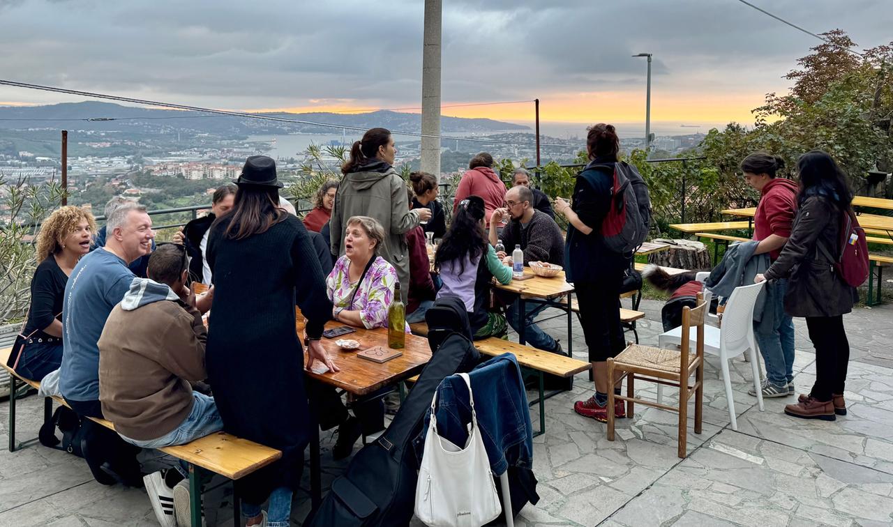 Gesellige Runde von Menschen an langen Holztischen in einer Buschenschank mit Blick auf Triest bei Sonnenuntergang.