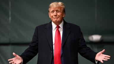 Donald Trump steht vor Videowand mit der Einblendng "Trump wins Iowa".