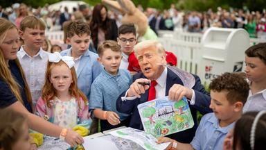 Ein älterer Mann im Anzug sitzt an einem Tisch, umringt von Kindern und hält ein gemaltes Bild und einen Stift in den Händen.