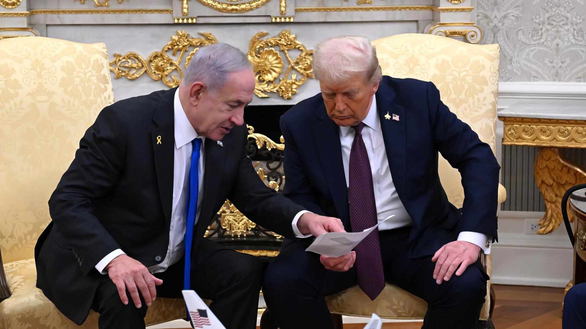 Zwei ältere Männer in dunklen Anzügen sitzen auf goldenen Stühlen und beugen sich über ein Blatt Papier, das einer in der Hand hält.