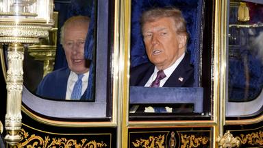 Zwei ältere Männer im Anzug sitzen in einer goldverzierten Kutsche und schauen heraus.