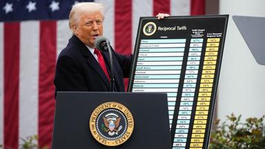 Ein älterer Mann im Anzug hält eine Tafel mit seinen Zöllen nach oben.