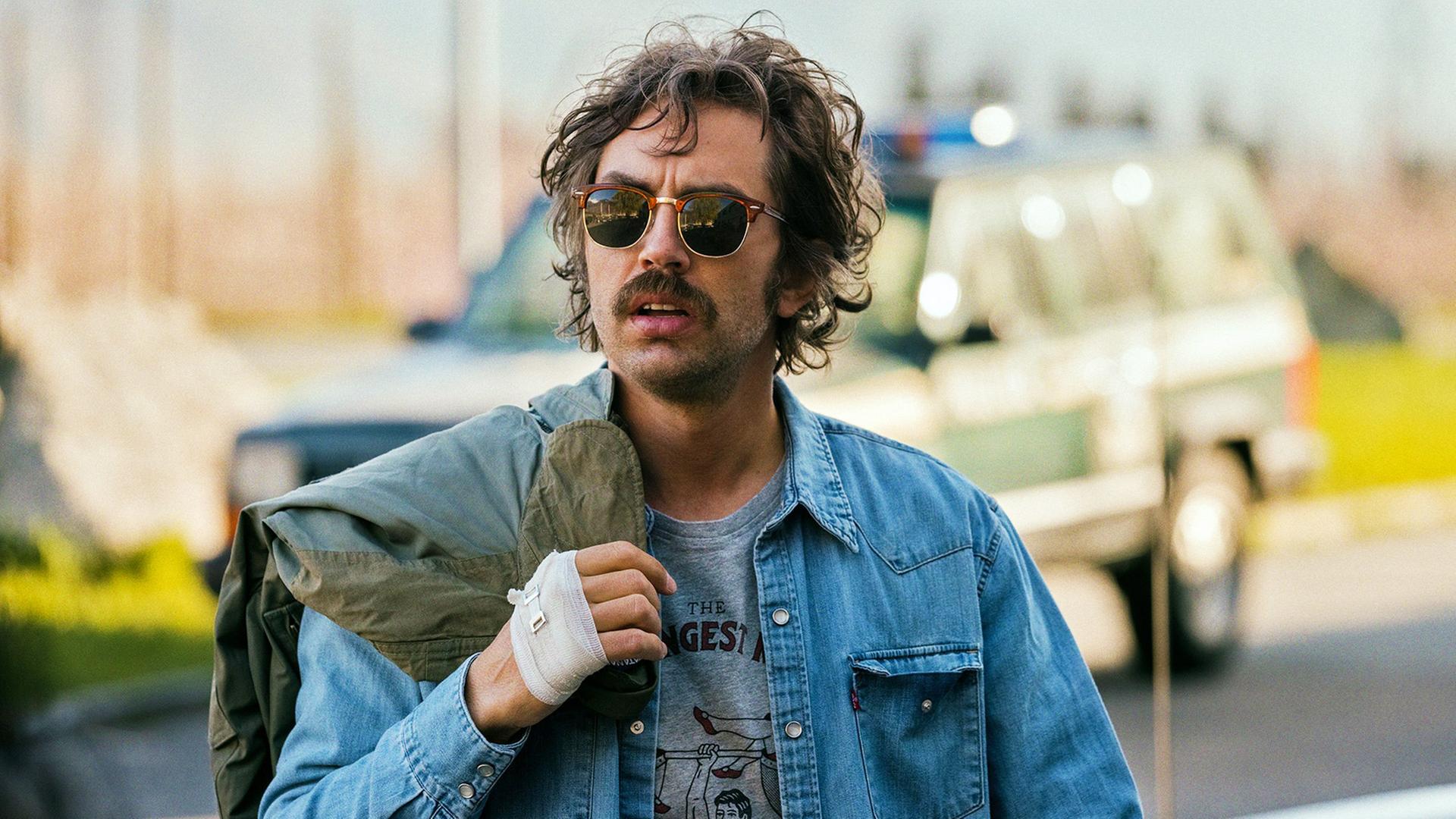 Ein Mann mit Schnurrbart und Sonnenbrille trägt eine Jeansjacke über einem T-Shirt mit Aufdruck. Er hat eine Jacke über die Schulter geworfen und eine bandagierte Hand. Im Hintergrund steht ein Polizeifahrzeug.