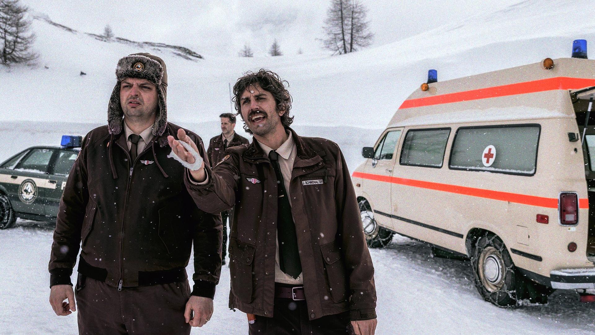 Zwei Männer in braunen Polizeiuniformen stehen im Schnee vor einem Rettungswagen; einer trägt eine Pelzmütze, der andere gestikuliert mit der Hand. Im Hintergrund weitere Beamte und Polizeifahrzeuge.