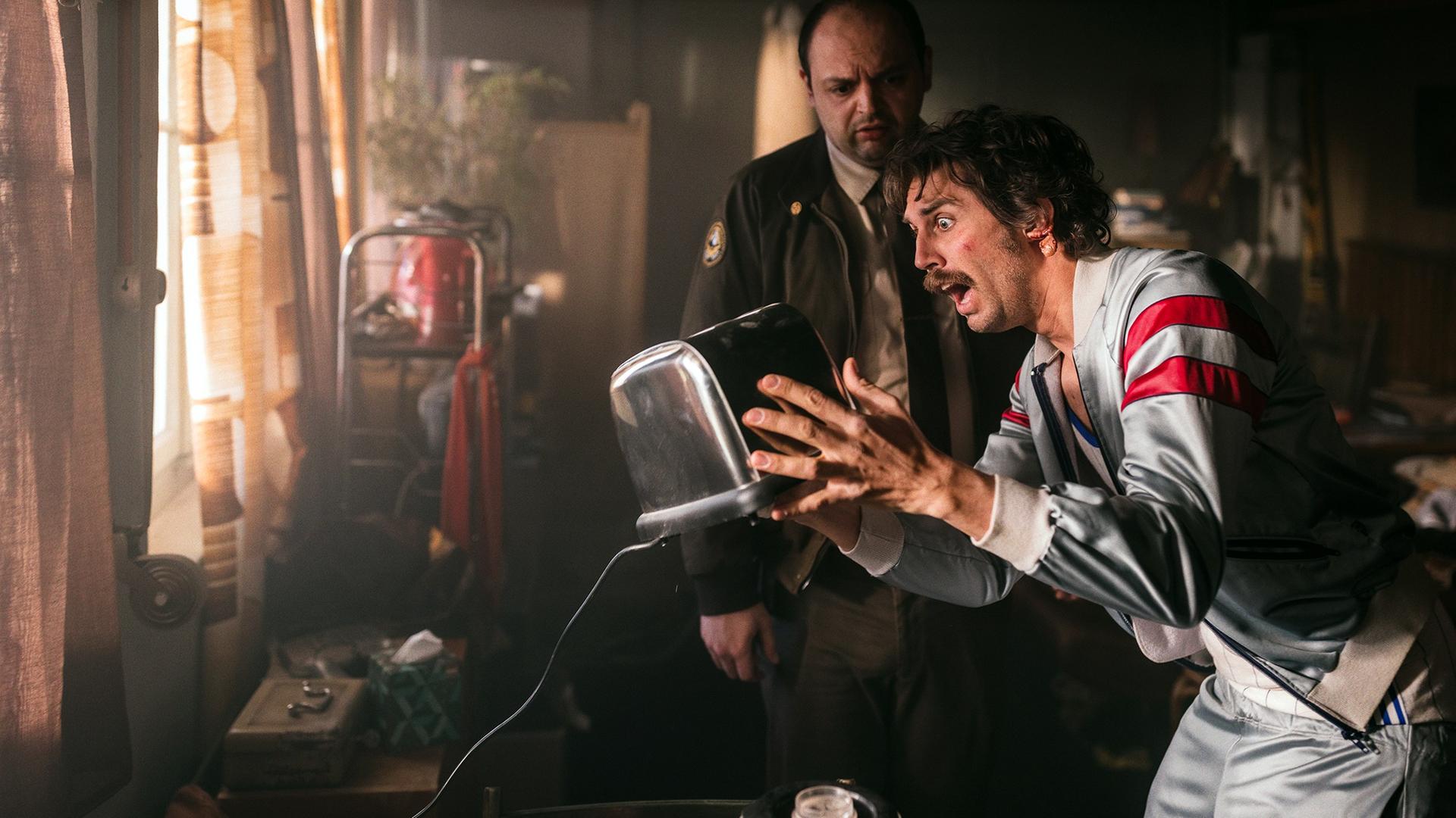 Zwei Männer in einem spärlich beleuchteten Raum; der eine im Trainingsanzug hält aufgeregt einen Toaster, der andere in Polizeiuniform blickt skeptisch darauf.