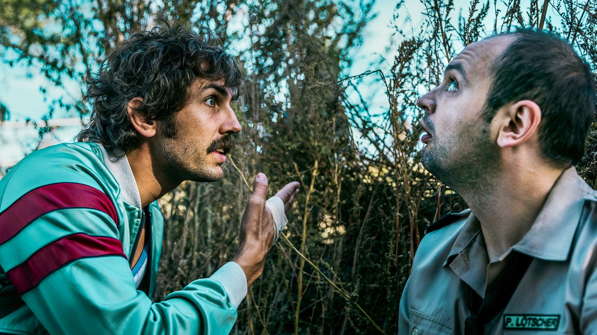 Zwei Männer kauern zwischen hohen, trockenen Pflanzen; der eine trägt eine türkis-rote Sportjacke und gestikuliert mit erhobener Hand, der andere in Uniform mit Namensschild blickt erschrocken nach oben.