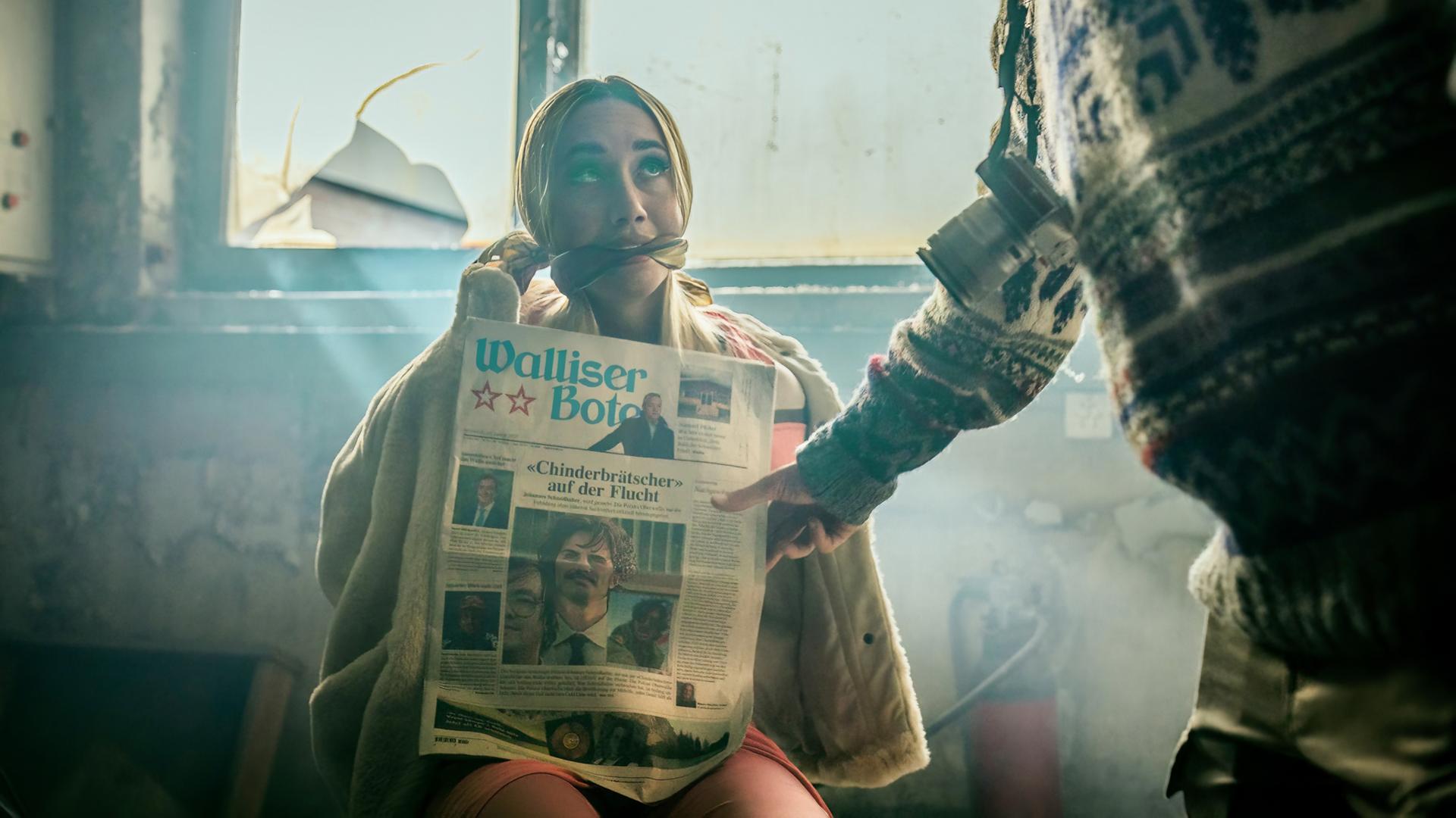 Annalena Miano als Valmira sitzt gefesselt und geknebelt in einem verlassenen Raum, hält eine aufgeschlagene Zeitung vor sich; Lichtstrahlen fallen durch ein zerbrochenes Fenster.