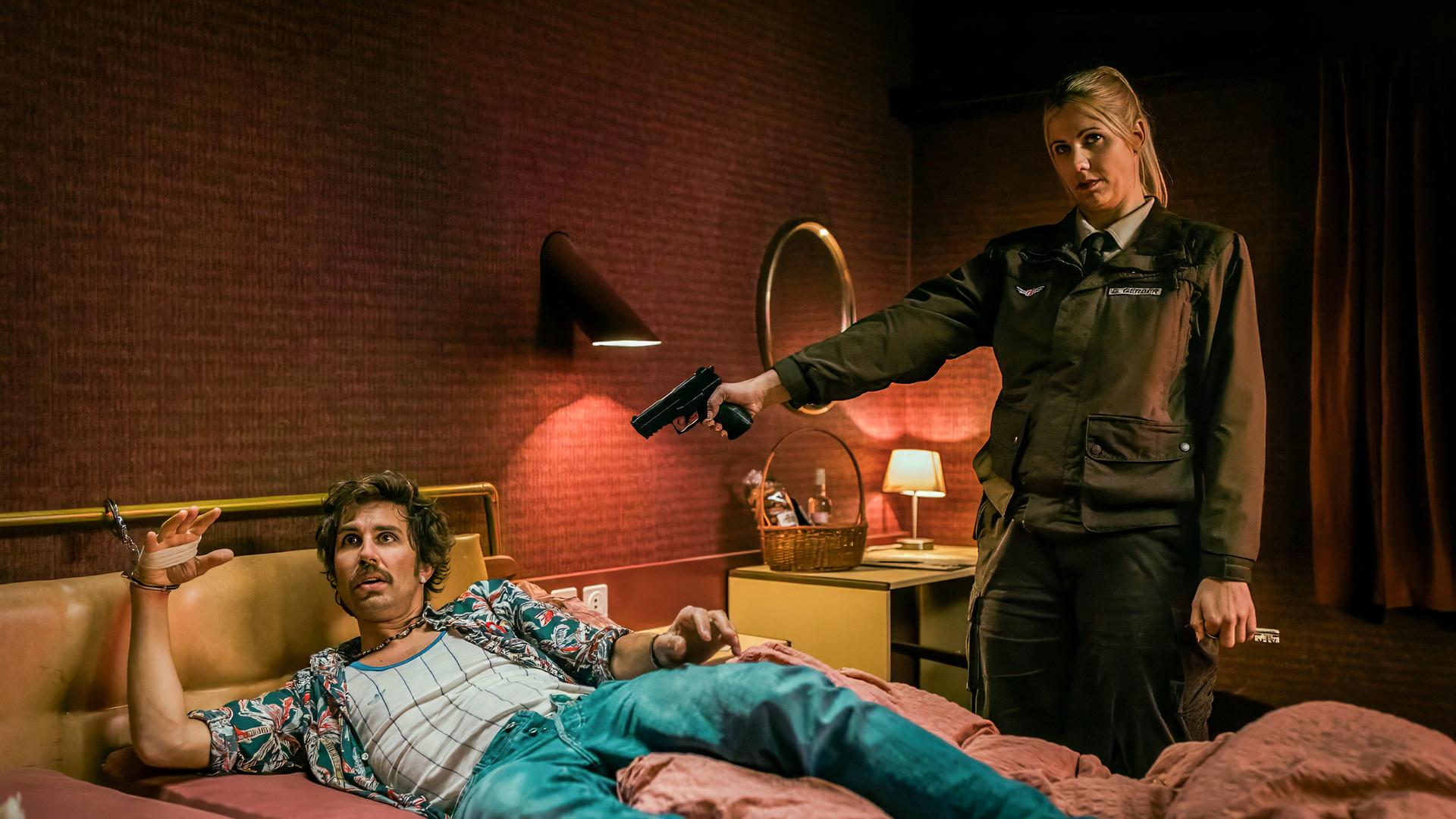 Ein Mann (David Constantin als Bax) liegt gefesselt auf einem Bett in einem düster eingerichteten Zimmer mit roter Tapete; Clelia Fux als Gerda steht daneben, richtet eine Pistole auf ihn und hält einen Schlüssel in der Hand.
