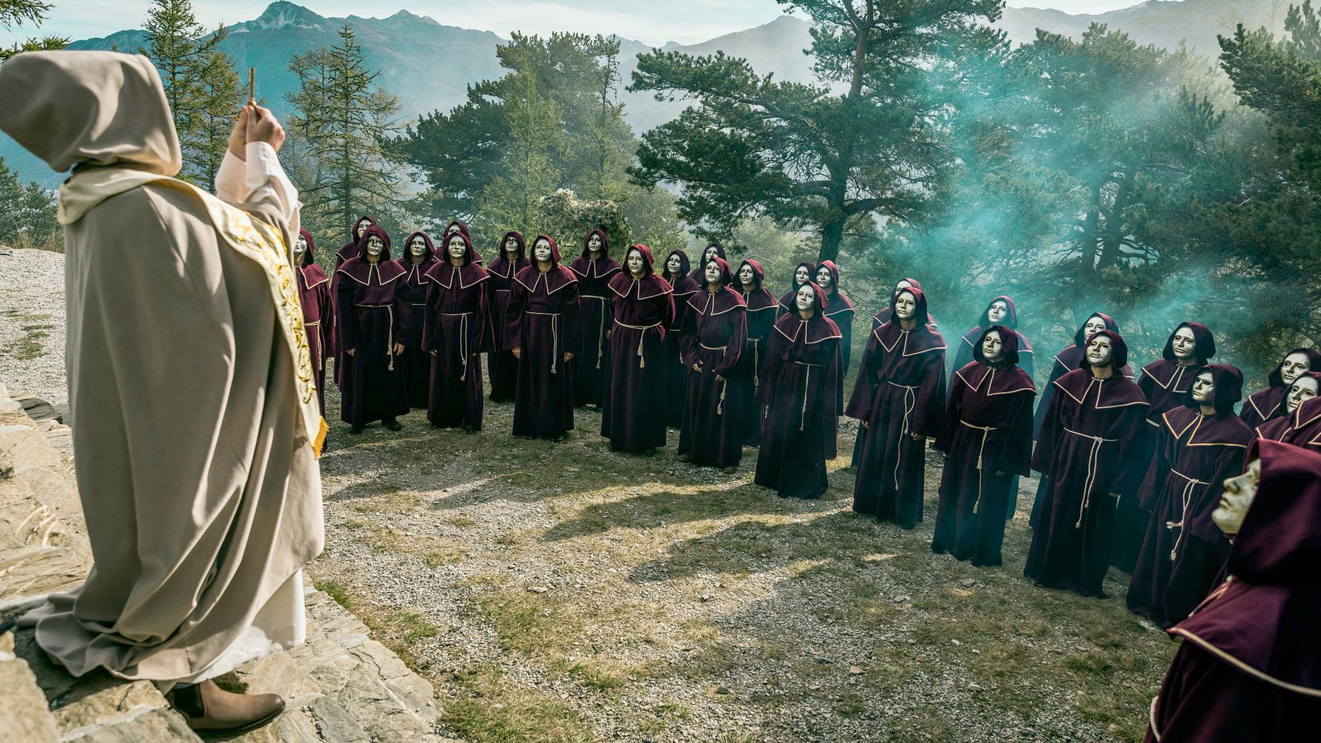 Eine harrende Menschenmenge in weinroten Kapuzenroben und goldenen Masken steht im Wald vor einem erhöhten, von hinten sichtbaren Gewandeten, der ein goldenes Kreuz in die Luft hält; im Hintergrund Berge und Bäume.