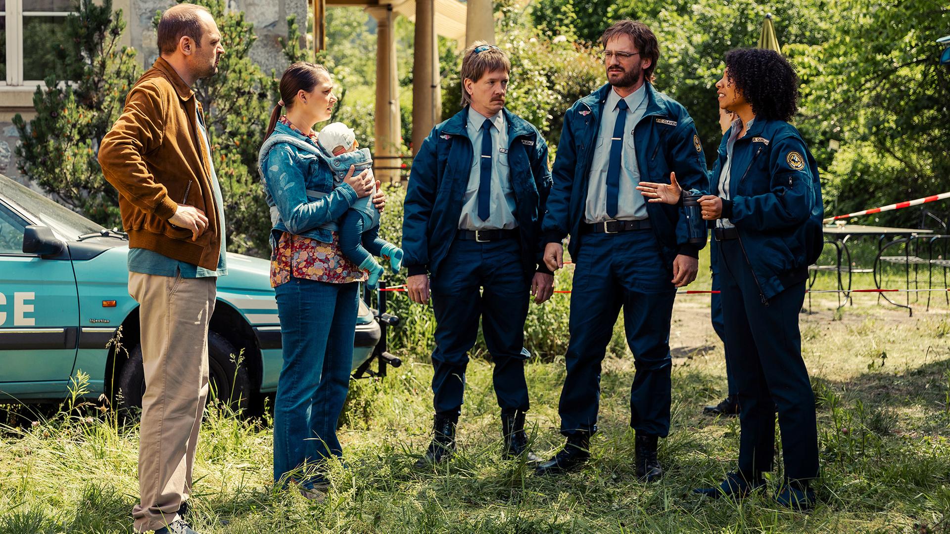 Fünf Personen stehen vor einem steinernen Haus und einem Polizeiwagen im Grünen: Dragan Vujic als Pirmin in brauner Jacke, Lena Furrer als Regina mit einem Baby im Arm, Vincent Veillon und Vincent Kucholl als uniformierte Commissaires Jean-Claude und Jean-Pierre sowie Safi Martin Yé als Polizeichefin im Gespräch.