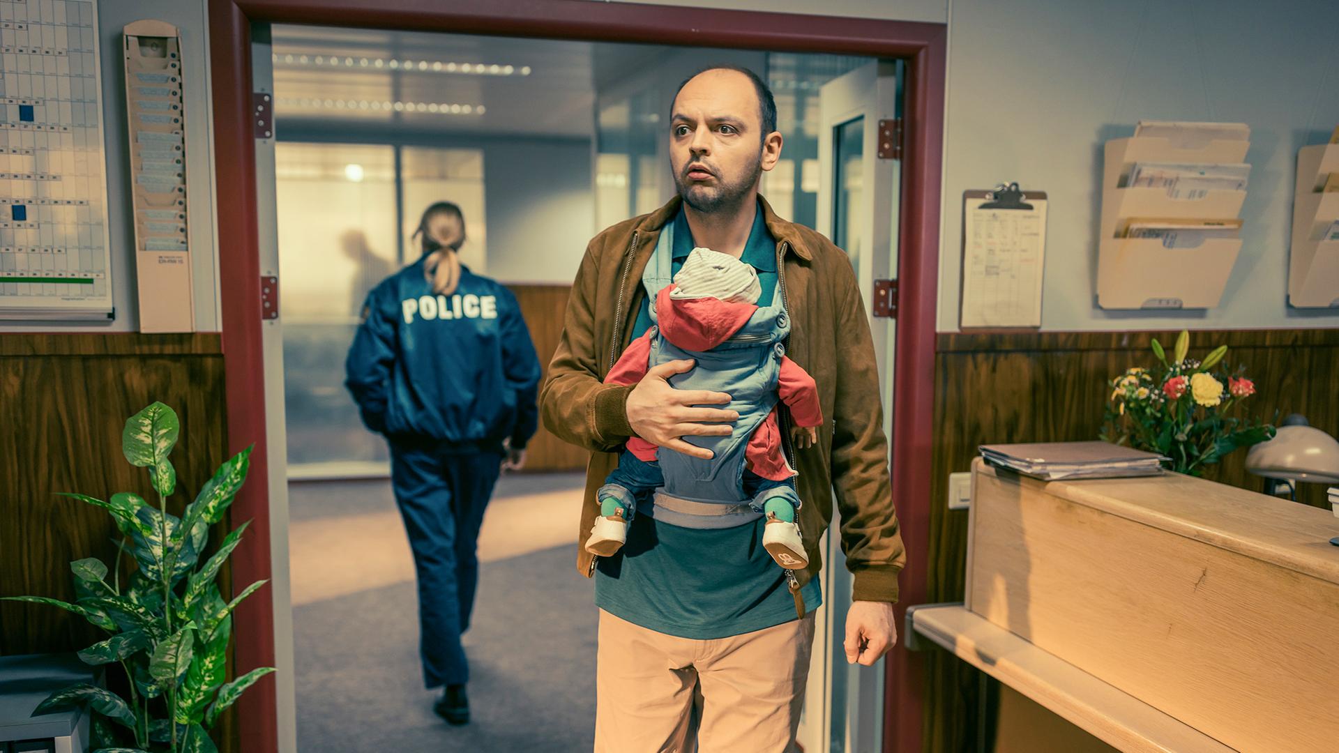 Dragan Vujic als Pirmin steht in einer Polizeiwache und trägt ein Baby in einer Bauchtrage; im Hintergrund verlässt eine Beamtin mit „Police“-Jacke den Raum, während Pirmin unsicher in Richtung Kamera blickt.