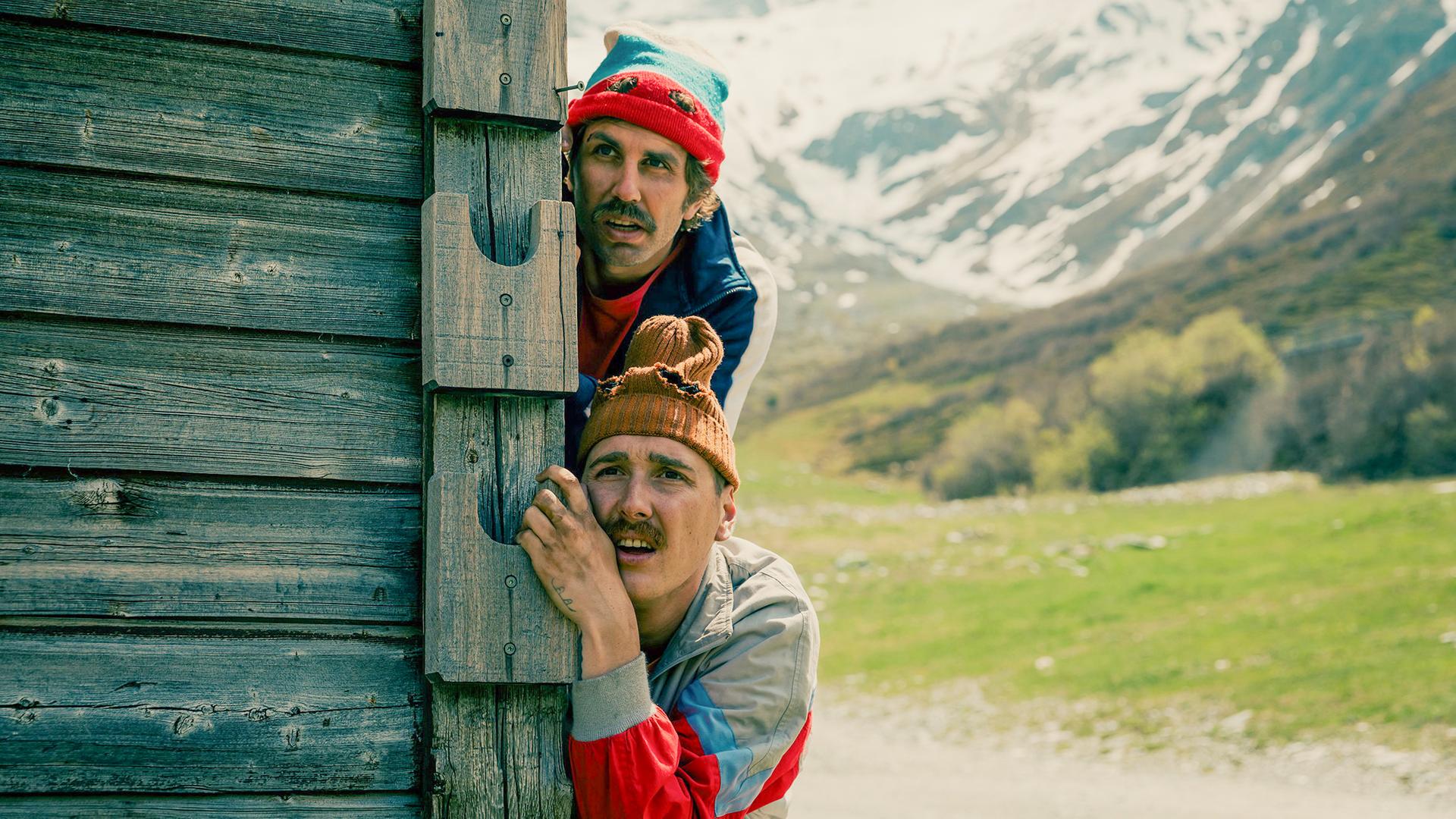 Zwei Männer kauern hinter einer Holzhütte in den Bergen; der obere trägt eine rote Mütze und blickt suchend über den Kopf des anderen, beide wirken angespannt.