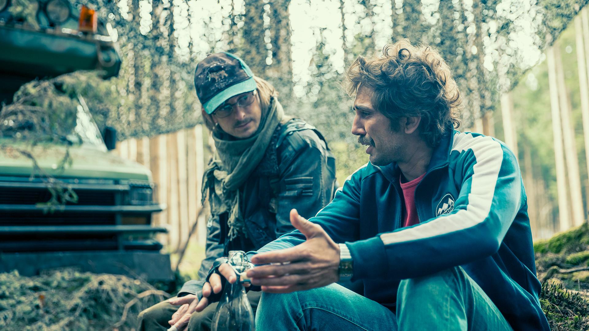 Cedric Schild als Smetterling und David Constantin als Bax sitzen unter einem Tarnnetz in einem Wald; Smetterling blickt Bax an, während Bax mit ausladender Geste gestikuliert.