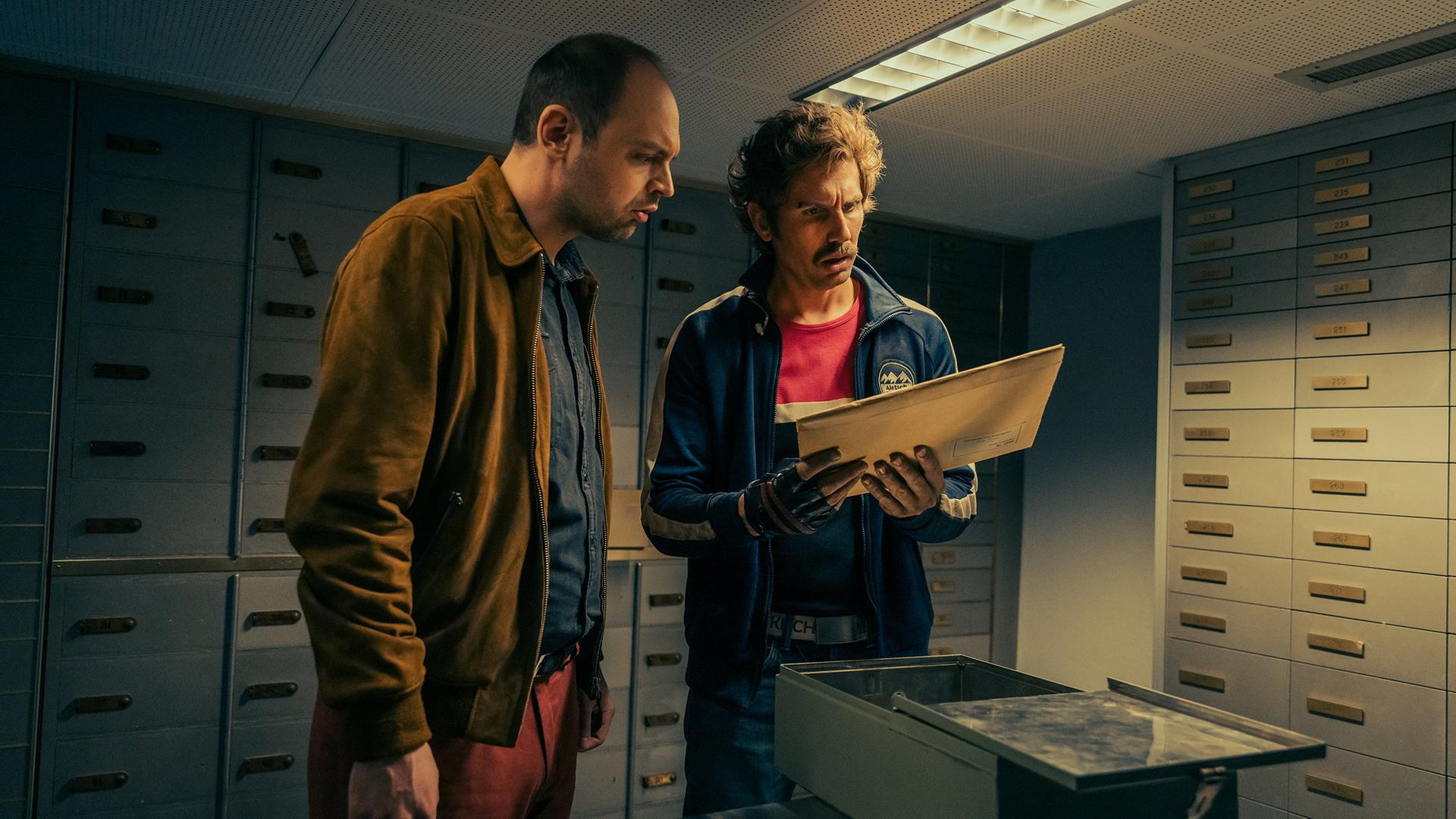 Dragan Vujic als Pirmin (links) und David Constantin als Bax (rechts) stehen in einem Schließfachraum vor metallenen Fachschränken; sie halten einen geöffneten Umschlag und betrachten gespannt die darin enthaltenen Dokumente.