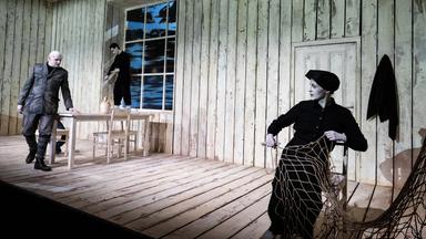 Theaterszene in einer hölzernen Hütte mit drei Schauspielenden. Links geht ein älterer Mann in einer Militäruniform am Tisch vorbei. Ein jüngerer Mann in schwarzer Kleidung steht barfuß auf dem Fensterbrett und schnitzt. Rechts im Vordergrund sitzt eine Frau in schwarzer Kleidung mit Kopfbedeckung auf einem Stuhl und arbeitet an einem großen Fischernetz. 