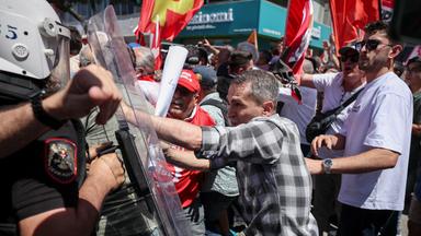 Bei einer Demonstration schiebt ein Mann gegen den Plexiglasschild eines Polizisten in Schutzausrüstung. Hinter ihm schweken Männer rote Fahnen.