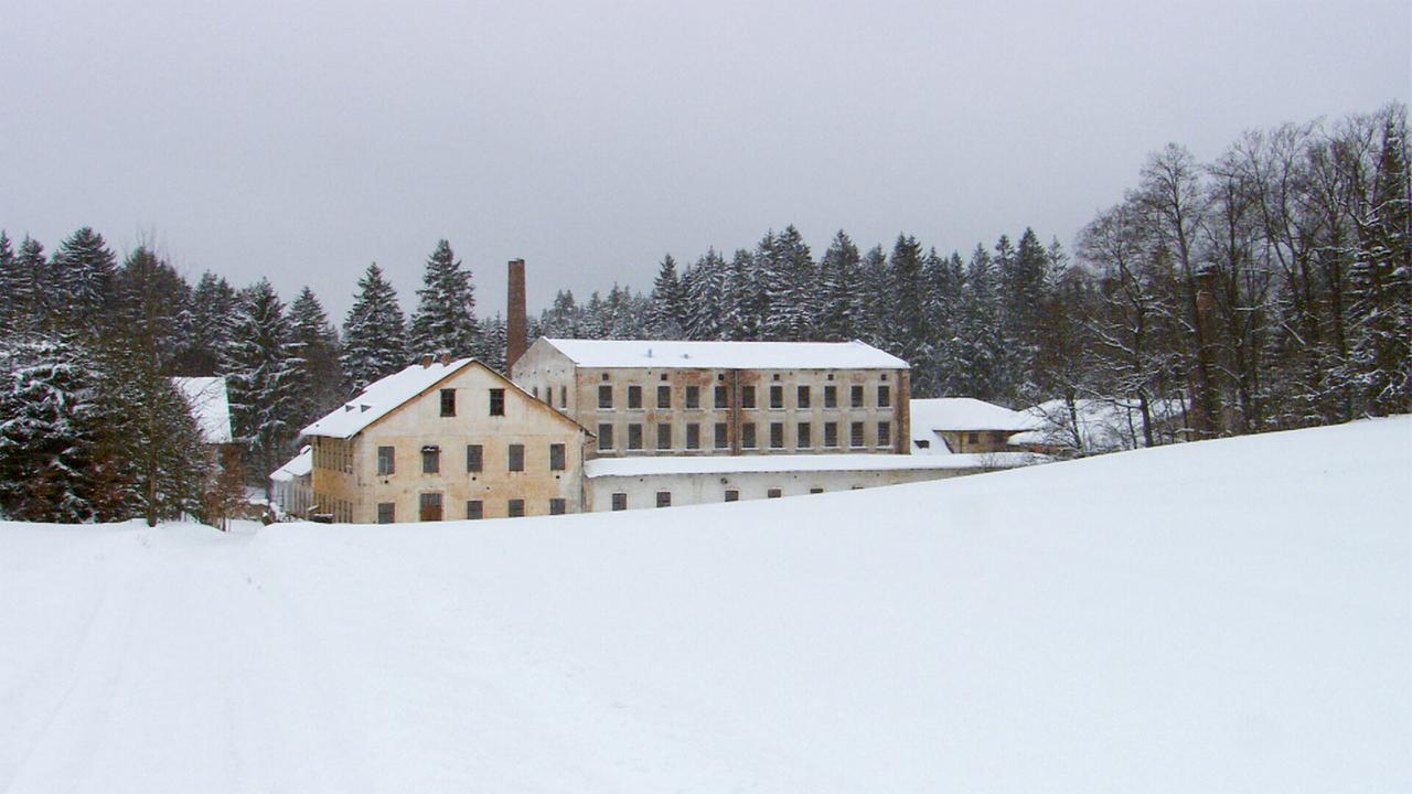 Das Bild zeigt ein winterliches Landschaftsbild mit einer alten Textilfabrik. Die Fabrik besteht aus zwei Gebäuden, die teilweise verfallen sind, umgeben von schneebedeckter Fläche und Bäumen im Hintergrund.