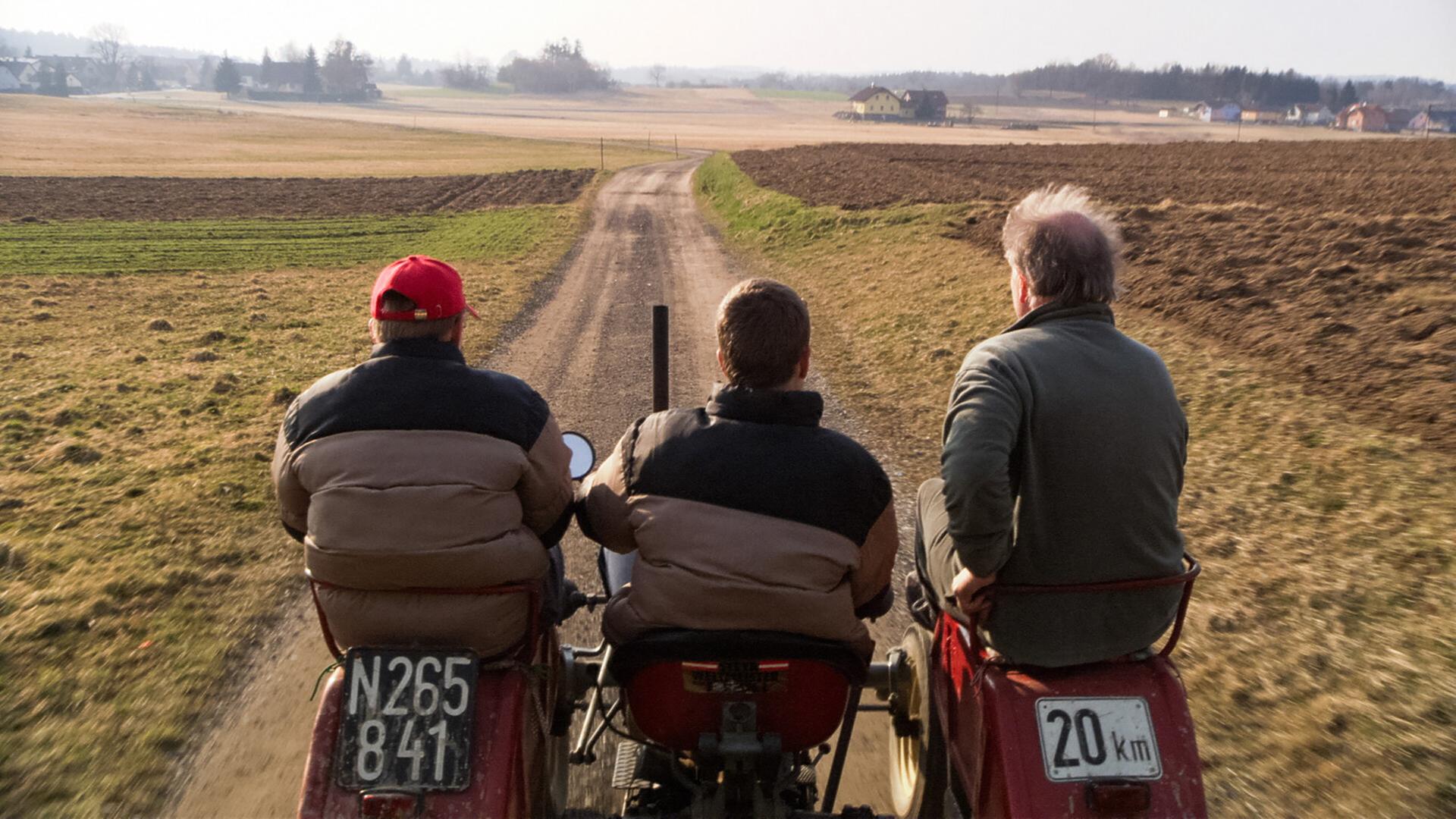 Drei Männer sitzen auf Traktoren und fahren einen unbefestigten Weg entlang. Im Hintergrund ist eine weite Landschaft mit Feldern und gelegentlichen Bäumen zu sehen.