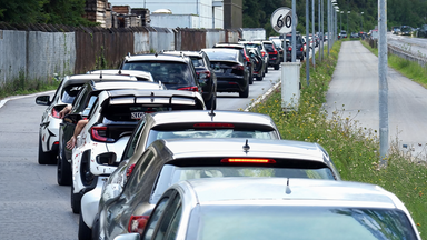 Langer Rückstau von Autos auf einer Straße neben einem Industriegelände. Einige Fahrer strecken ihre Arme aus den Autofenstern, während die Fahrzeuge dicht an dicht stehen. Rechts neben der Straße verläuft ein leerer Gehweg, gesäumt von Straßenlaternen und Wildblumen.