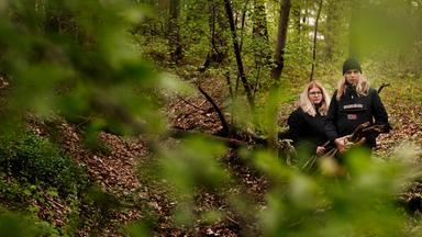 Teilweise hinter grünem Laub verdeckt, stehen Emely und Pauline im Wald. Sie tragen mehrere Äste in den Händen.