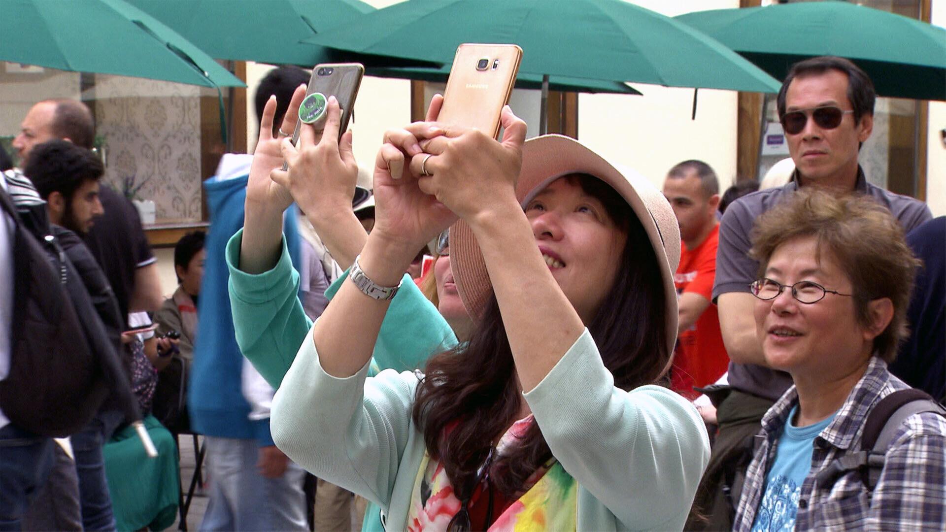 Das Bild zeigt eine belebte Straßenszene mit mehreren Touristen, die ihre Smartphones in die Höhe halten, um Fotos zu machen. Im Vordergrund ist eine Frau mit langen, dunklen Haaren und einem breiten, hellen Hut zu sehen. Sie lächelt, während sie mit beiden Händen das Smartphone hält. Ihre Kleidung ist hellblau und kunterbunt. Hinter ihr stehen weitere Personen, darunter eine Frau mit Brille und einem karierten Hemd sowie ein Mann mit Sonnenbrille und einem neutralen Gesichtsausdruck.   Im Hintergrund sind einige Tische und Schirme zu sehen, die darauf hindeuten, dass es sich um ein Café oder ein Restaurant handeln könnte. Die gesamte Atmosphäre wirkt lebhaft und touristisch, mit einer Vielzahl von Menschen in der Szene. Die Umgebung ist urban und scheint ein beliebtes Ziel für Touristen zu sein.