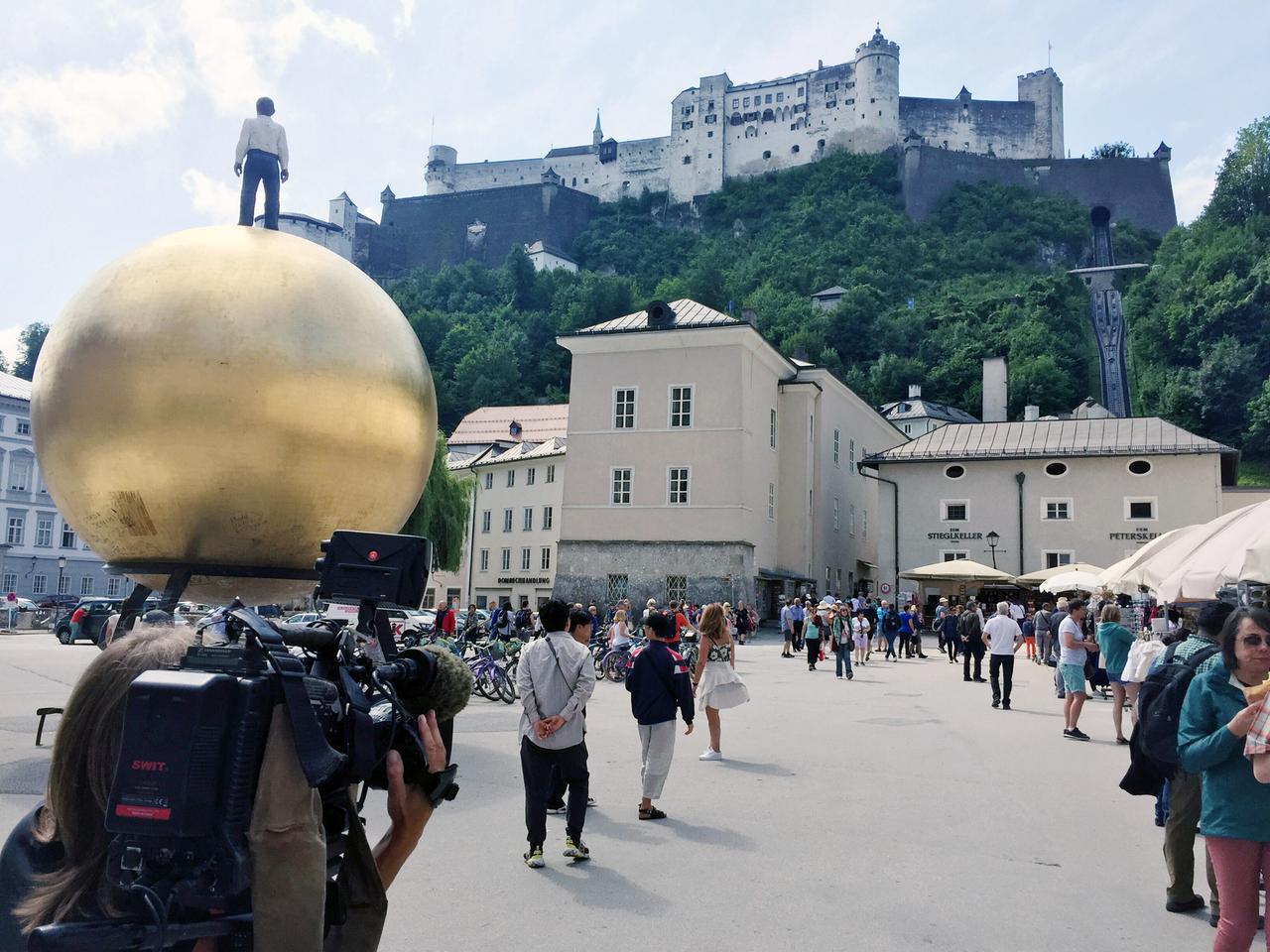 Das Bild zeigt eine belebte Szenerie in der Salzburger Altstadt. Im Vordergrund steht eine große, goldene Kugel, auf der eine kleine Figur steht. Die Kugel ist glänzend und rund, während die Figur in moderner Kleidung dargestellt ist.   Links im Bild sieht man eine Gruppe von Menschen, die sich unterhalten und sich in die Richtung der Kamera bewegen. Hinter ihnen sind Fahrräder abgestellt. Auf der rechten Seite befinden sich mehrere Tische mit Sonnenschirmen, die auf eine Gastronomie hinweisen.   Im Hintergrund erhebt sich die imposante Festung Hohensalzburg auf einem Hügel, mit mehreren Türmen und einer Mauer, die sich entlang der Bergkuppe zieht. Der Himmel ist teilweise bewölkt, und es herrscht eine lebhafte Atmosphäre, die den Tourismus in der Stadt widerspiegelt.   Die Szene vermittelt ein Gefühl von geschäftigem Treiben und der Konkurrenz zwischen den touristischen Attraktionen und den Bedürfnissen der ansässigen Bevölkerung.