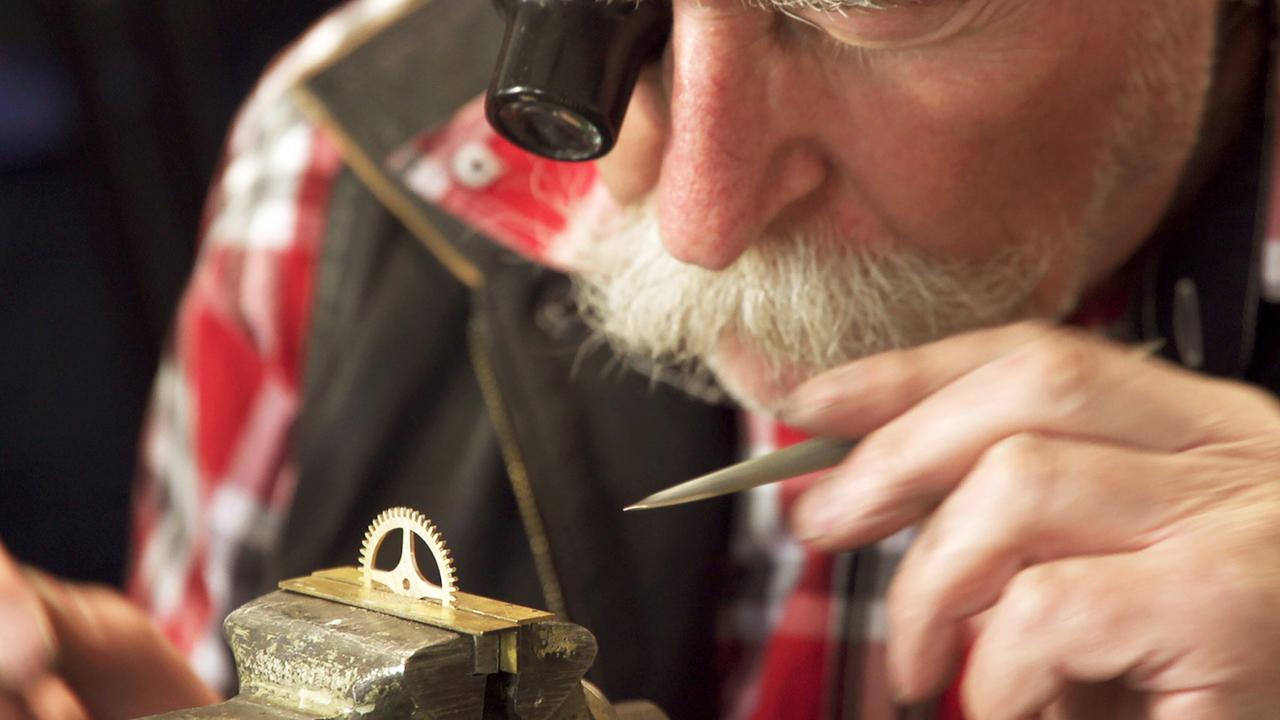 Das Bild zeigt einen älteren Mann, der konzentriert an einem Uhrwerk arbeitet. Er trägt eine Brille und hat einen weißen Schnurrbart. Der Mann beugt sich über eine Werkbank, auf der ein kleines Zahnrad aus hellem Metall liegt. Es hat mehrere Zähne und wird von einem halben Kreis umrahmt. Seine linke Hand hält ein Werkzeug, während die rechte Hand ruhig über dem Zahnrad schwebt, als ob er präzise arbeiten würde. Im Hintergrund sind unscharfe Werkzeuge und Materialien zu erkennen, die auf eine Werkstatt hinweisen. Die Farbgestaltung des Bildes wirkt natürlich, mit erdigen Tönen und einem Hauch von Rot in seinem karierten Hemd, das er unter einer Weste trägt. Das Bild vermittelt eine Atmosphäre von Handwerkskunst und Konzentration.