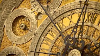 Das Bild zeigt eine Detailansicht eines kunstvoll gestalteten Zifferblattes einer Uhr. Der Hintergrund ist in einem warmen Goldton gehalten, während die Ziffern und Zeiger aus poliertem Metall bestehen.   Im Zentrum der Uhr ist ein rundes Ziffernblatt mit filigranen Verzierungen zu sehen. Die Ziffern sind auf einem kreisförmigen Bereich aufgetragen, umgeben von dekorativen Elementen. Einige der Ziffern stehen in römischer Zahl, was auf einen klassischen Stil hinweist.  Die Uhr hat mehrere kleine Ziffernblätter, die für verschiedene Funktionen stehen, wie beispielsweise eine Datumsanzeige. Diese sind ebenfalls detailliert und mit feinen Mustern verziert. Der Stundenzeiger und der Minutenzeiger sind auffällig, wobei der Minutenzeiger länger ist und eine geschwungene Form hat.   Die gesamte Komposition vermittelt den Eindruck von Handwerkskunst und präziser Technik, was auf die Tradition des Uhrmacherhandwerks hinweist, die im Bild beschrieben wird.