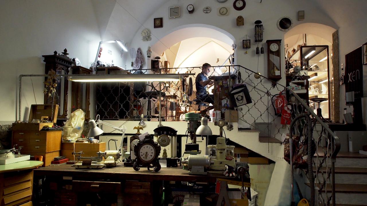 Das Bild zeigt eine Uhrmacherwerkstatt mit einer klar strukturierten, aber vollgepackten Arbeitsumgebung. Im Vordergrund befindet sich ein großer Tisch, auf dem mehrere Uhren, Werkzeuge und Maschinen zu sehen sind. Als zentrale Objekte sind eine große, antike Uhr, eine Tischleuchte und kleinere Uhrenteile hervorgehoben.   Im Hintergrund erkennt man einen Handwerker, der an einem Tisch sitzt und an einer Uhr arbeitet. Er trägt eine dunkle Kleidung und ist mit verschiedenen Werkzeugen umgeben, die auf einem weiteren Tisch platziert sind. An der Wand sind zahlreiche Uhren und Uhrenteile zur Dekoration oder als Ausstellung aufgehängt. Darüber hinaus gibt es einen Treppenaufgang, der zu einem höheren Bereich der Werkstatt führt.  Die Beleuchtung ist funktional und kommt von mehreren Lichtquellen, die die Werkstatt gut ausleuchten. Insgesamt vermittelt das Bild einen Eindruck von Tradition und Handwerkskunst im Bereich der Uhrmacherei.