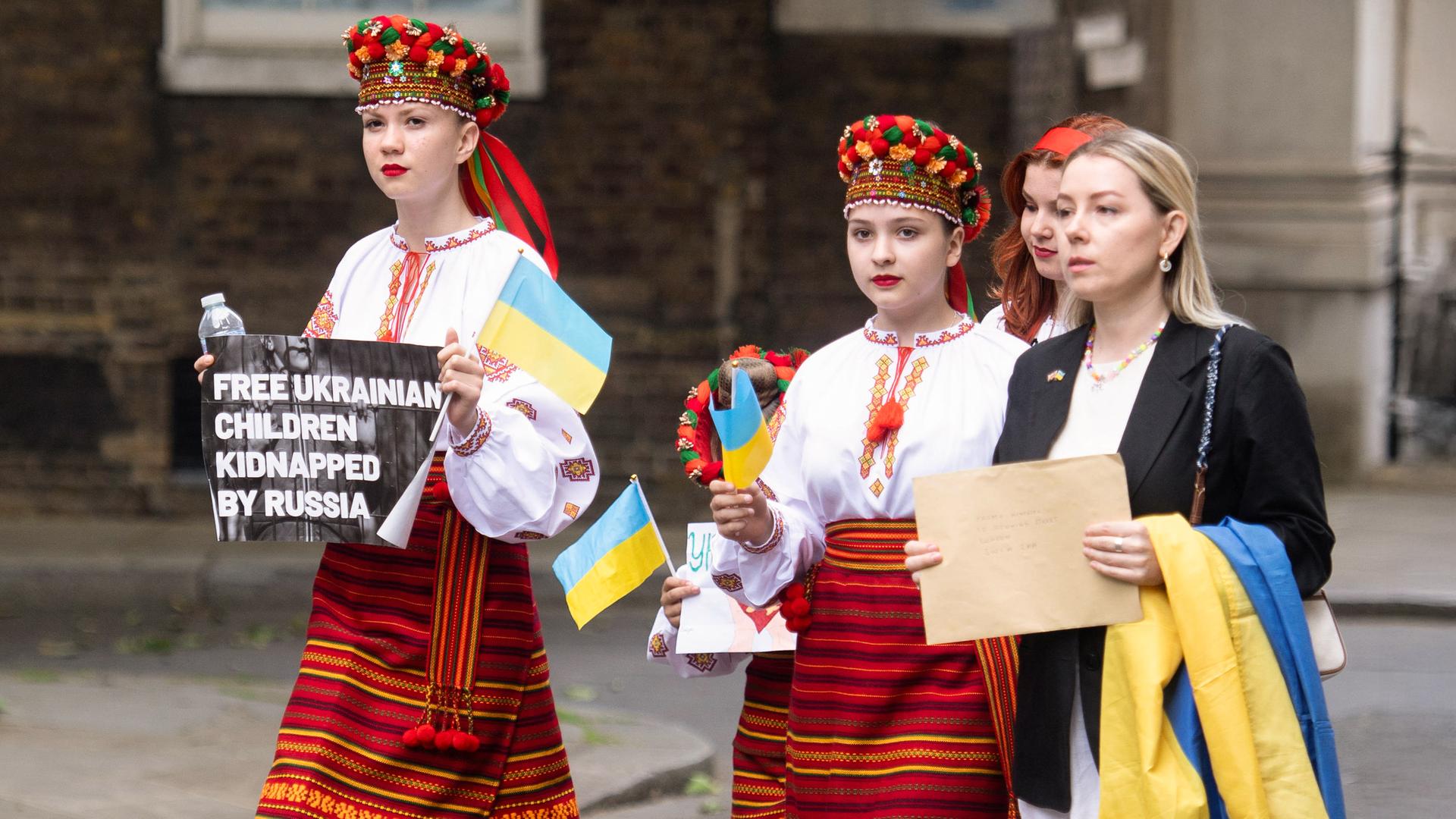 Drei junge Mädchen in ukrainischer Tracht und ukrainischen Fahnen in der Hand laufen über die Straße. Ein Mädchen trägt ein Schild mit der Aufschrift: Free Ukrainian Children Kidnapped by Russia. Mit dabei ist eine Frau, die einen braunen Umschlag in der Hand hält.