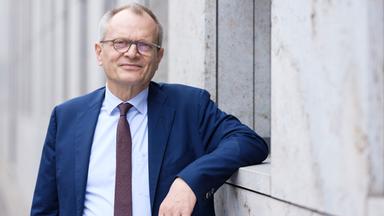 Ein Mann mit Brille und Anzug lehnt an einem grauen Gebäude.